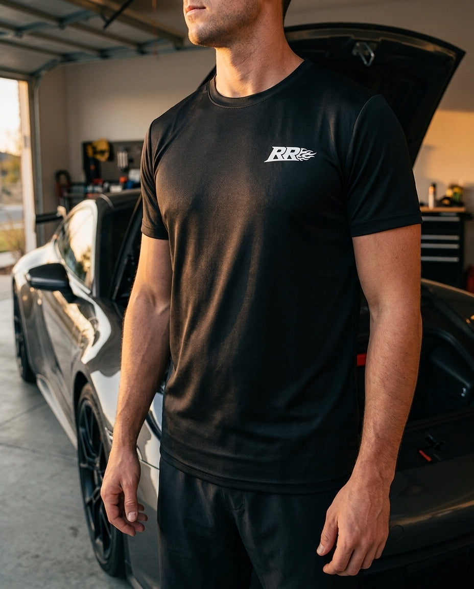 Rowdy Ridez Performance Tee worn beside an open-hood sports car in a garage at dusk